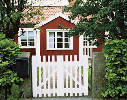 Röd träbyggnad med vita fönster och dörr, framför en vit trädgårdsgrind med gröna buskar runt.