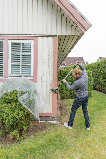 En person tvättar träpanel på ett hus med en borste, nära gröna buskar.