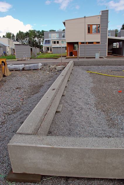 Betongfundament framför ett hus under byggnation, med grusmark i förgrunden och byggnadsmaterial i bakgrunden.