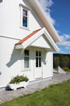 Vitt hus med rött tegeltak, vit dörr med glasrutor, blomsterrabatt framför, grå stengång och gräsmatta.