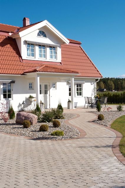 Vit villa med rött tak och vita fönster. Stenlagd gångväg leder till entrén. Buskar och trädgårdsmöbler syns i förgrunden.