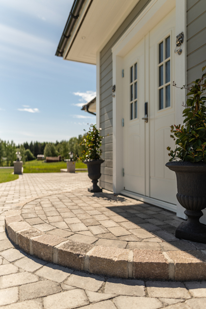 Entré med stenbelagd gång, två krukväxter vid dörren. Soligt väder, gräsmatta och skog i bakgrunden.