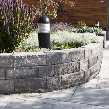 Stenlagd mur med omgivande växter och en pollare i trädgårdslandskap.