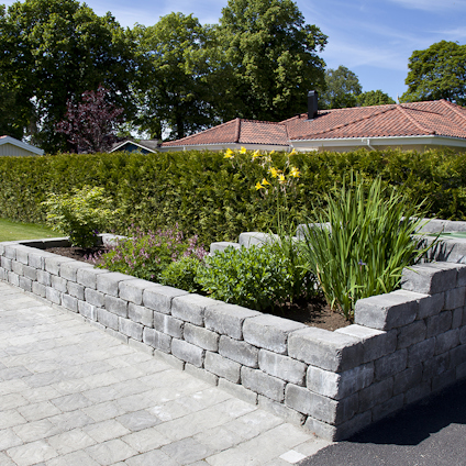 Stenmur av rektangulära block med planterad växtlighet ovanpå, mot en bakgrund av träd och ett rött hustak.