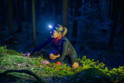 Person med pannlampa kryper i mörk skog, omgiven av mossa och vegetation.