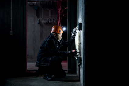 Person i orange hjälm och pannlampa arbetar med elinstallationer i ett mörkt rum.