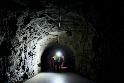 Två personer med hjälm och ficklampa går i en mörk tunneltunnel med rå bergvägg.