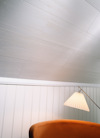 Vitt träpaneltak och vägg med vita lodräta paneler; en golvlampa med vit skärm vid en brun soffa.
