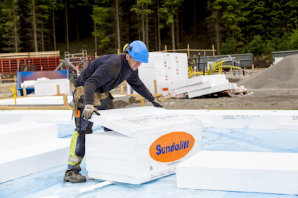 Man i blå hjälm hanterar en vit Sundolitt isoleringsskiva på en byggarbetsplats.