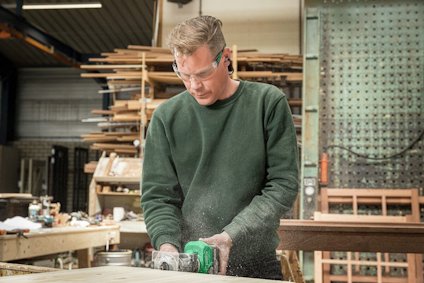 En person med skyddsglasögon sliper en trädskiva inomhus. Trähyllor och arbetsbänkar i bakgrunden.