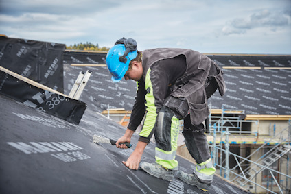 Person i blå hjälm installerar takmaterial på sluttande tak med utsikt över ställningar och molnig himmel.